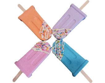 May include: Four popsicle-shaped objects arranged in an X pattern. Each features a different pastel color: orange, lavender, pink, and blue. Each has a wooden stick at the base and a white textured topping decorated with colorful sprinkles.