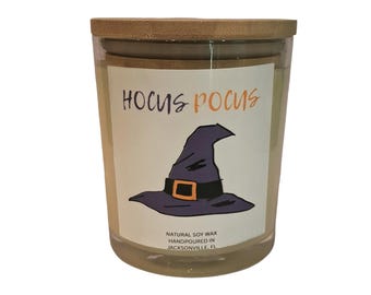 May include: A clear glass candle jar with a bamboo lid featuring a white label with the text HOCUS POCUS in purple and orange, a purple witch hat illustration, and the text NATURAL SOY WAX HANDPOURED IN JACKSONVILLE, FL at the bottom.