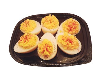 May include: Six halved hard-boiled eggs arranged on a black plastic tray. Each egg white half is filled with a piped, bright yellow yolk mixture, topped with a light dusting of red paprika seasoning, creating a high-contrast food presentation.