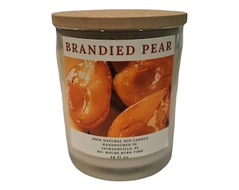 May include: A clear glass candle jar with a light wood lid, featuring a label that reads BRANDIED PEAR with an image of sliced pears. The label text includes 100% NATURAL SOY CANDLE, HANDPOURED IN JACKSONVILLE, FL, 60+ HOURS BURN TIME, and 14 fl oz.