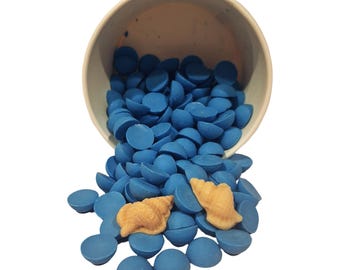 May include: A light-colored paper cup tipped over, spilling a large quantity of small, round, blue wax melt beads. Two light tan, textured seashell-shaped wax melts are resting on top of the pile of blue beads against a plain white background.