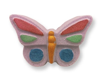 May include: A butterfly-shaped bath bomb in shades of lavender, red, green, blue, and gold. The wings have red and green accents, with blue circles on the lower wings. The body is gold, and the overall design is whimsical.