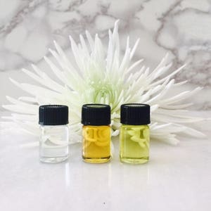 May include: Three small glass bottles with black caps, containing clear, yellow, and green liquids. The bottles are arranged in a row on a white marble surface, with a white flower behind them.