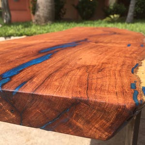 May include: A wooden bench with a live edge top and blue epoxy resin filling the cracks. The bench has a natural wood finish and is supported by two black metal legs.