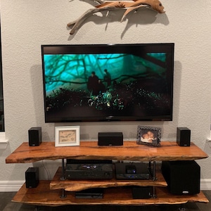 May include: A wooden entertainment center with a flat screen TV, speakers, and a receiver. The TV is playing a movie with a dark forest scene. The entertainment center has two shelves and is made of natural wood with a live edge.