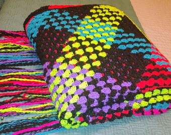 May include: A colorful crocheted blanket with a black background and fringe. The blanket is made with different colored yarn, including purple, green, yellow, blue, and red.