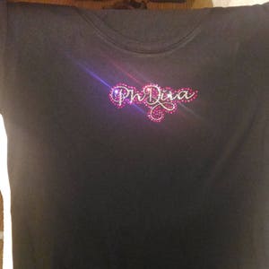 Peut inclure: T-shirt noir avec les mots "Mr. Diva" en lettres argentées pailletées avec des strass roses.