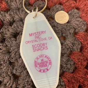 May include: A white plastic motel key chain with a purple inscription that reads "Mystery Inc. Crystal Cove, CA Scooby Snacks" and a purple circle with the text "Drop in any mailbox We Guarantee Postage".