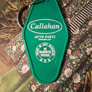 May include: Green vintage-style keychain with the text "Callahan Auto Parts" in white. The keychain has a metal ring and additional text that reads "Drop in any mailbox, we guarantee postage."