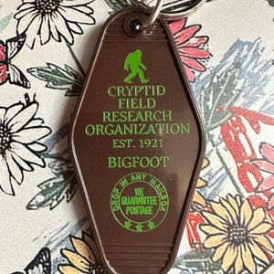 May include: Brown motel key chain with a green silhouette of Bigfoot and the text "Cryptid Field Research Organization Est. 1921 Bigfoot". The keychain also has a green circle with the text "Drop in any mailbox We Guarantee Postage" and three stars.