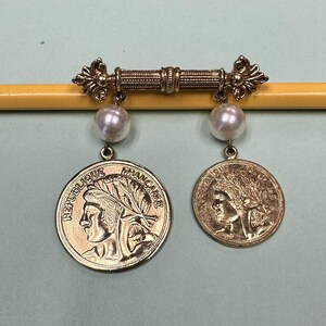 May include: A gold-toned metal brooch featuring a horizontal bar with decorative ends. Two white spherical beads hang from the bar, each attached to a circular gold-toned coin charm. The larger coin displays the text REPUBLIQUE FRANCAISE.