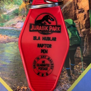 May include: Red plastic key chain shaped like a motel key with a black Jurassic Park logo, Isla Nublar, Raptor Pen text, and a black circle with the text "Drop in any mailbox, We Guarantee Postage".