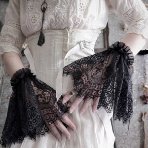 May include: A pair of black lace wrist cuffs with frilled edges. The cuffs are worn over a white, lace-trimmed blouse with a white corset.