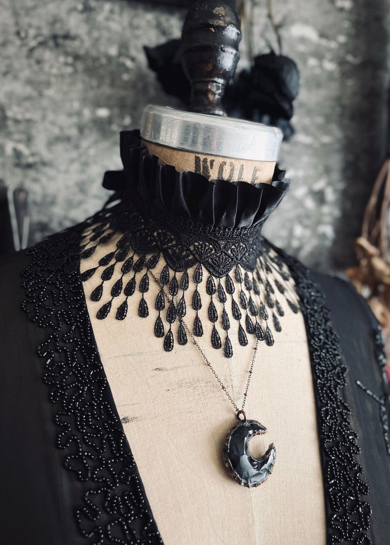 A wide black lace choker collar encircles the neck. Lace is detailed, textured and falls in dainty teardrop fringe past the collarbones. Choker is topped with wide black satin pleated trim. Tags romantic gothic dark fashion vampire wedding mourning