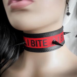 May include: Black choker with red ribbon featuring the text "I BITE" in bold, black letters. The choker has black spikes and a black leather-like band. A star-shaped earring hangs from the ear.