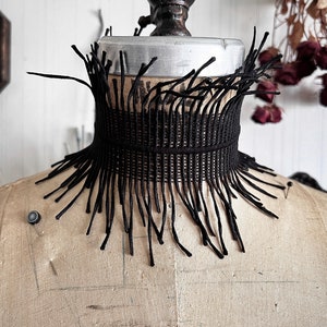 A wide black lace victorian choker necklace sits high on the neck. It has long feathered fringe emerging from both the top and bottom edges, framing the collarbones and face. The style can be described as avant garde and brutalist, or even costume.