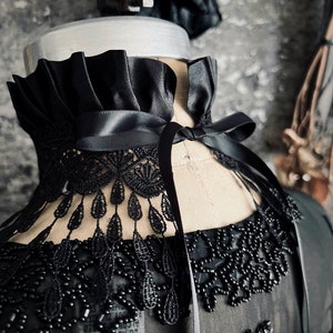 A wide black lace choker collar encircles the neck. Lace is detailed, textured and falls in dainty teardrop fringe past the collarbones. Choker is topped with wide black satin pleated trim. Necklace ties in back with wide black satin ribbon.