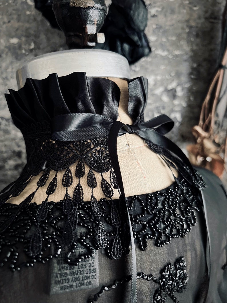 A wide black lace choker collar encircles the neck. Lace is detailed, textured and falls in dainty teardrop fringe past the collarbones. Choker is topped with wide black satin pleated trim. Necklace ties in back with wide black satin ribbon.