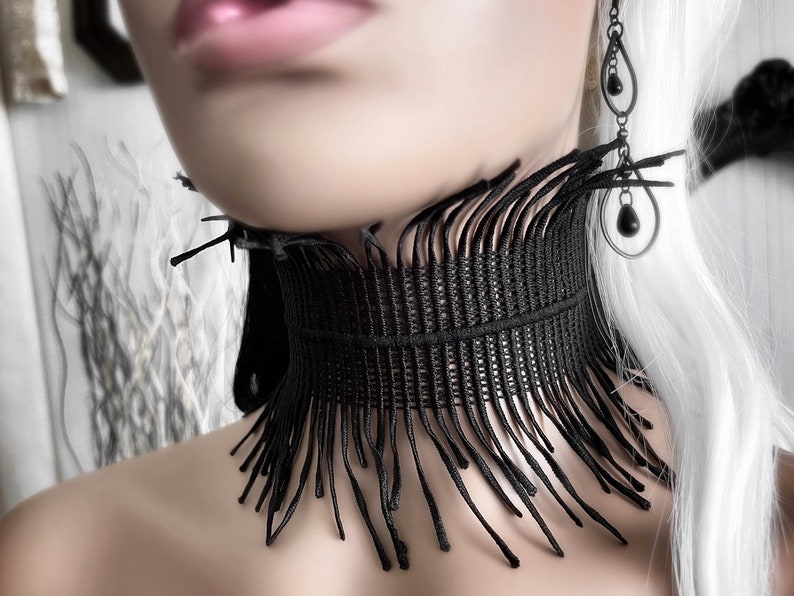 A wide black lace victorian choker necklace sits high on the neck. It has long feathered fringe emerging from both the top and bottom edges, framing the collarbones and face. The style can be described as avant garde and brutalist, or even costume.