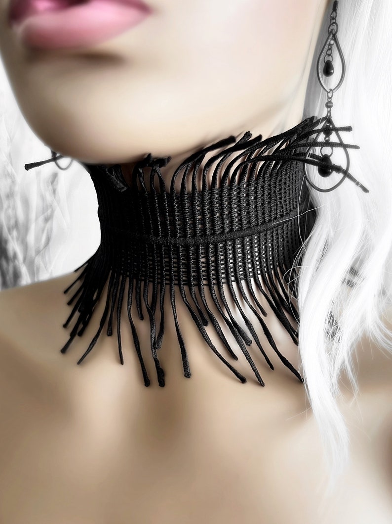 A wide black lace victorian choker necklace sits high on the neck. It has long feathered fringe emerging from both the top and bottom edges, framing the collarbones and face. The style can be described as avant garde and brutalist, or even costume.