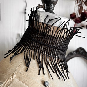 A wide black lace victorian choker necklace sits high on the neck. It has long feathered fringe emerging from both the top and bottom edges, framing the collarbones and face. The style can be described as avant garde and brutalist, or even costume.