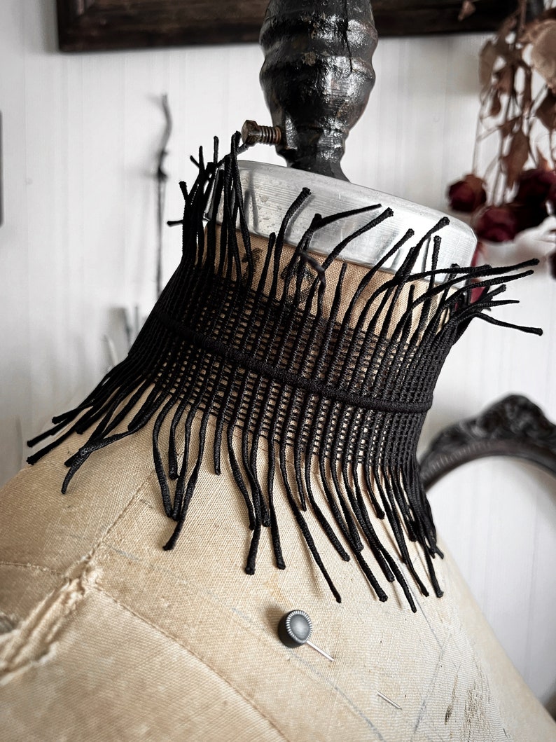 A wide black lace victorian choker necklace sits high on the neck. It has long feathered fringe emerging from both the top and bottom edges, framing the collarbones and face. The style can be described as avant garde and brutalist, or even costume.