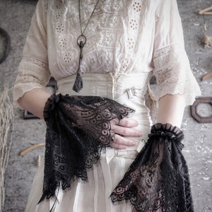 May include: A white lace blouse with a long, dangling key pendant necklace. The blouse has a fitted bodice and a full skirt. The model is wearing black lace wrist cuffs.