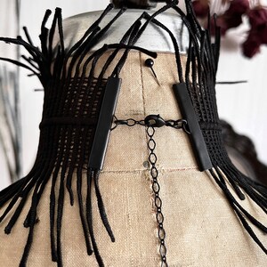 A wide black lace victorian choker necklace sits high on the neck. It has long feathered fringe emerging from both the top and bottom edges, framing the collarbones and face. The style can be described as avant garde and brutalist, or even costume.