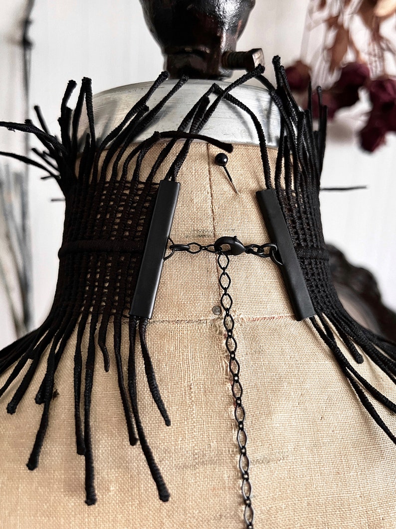 A wide black lace victorian choker necklace sits high on the neck. It has long feathered fringe emerging from both the top and bottom edges, framing the collarbones and face. The style can be described as avant garde and brutalist, or even costume.