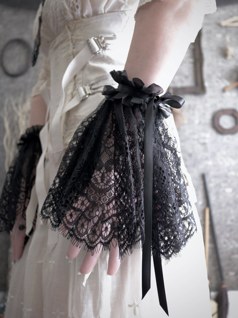 May include: Black lace wrist cuffs with a black satin ribbon. The cuffs are gathered at the wrist and have a delicate lace pattern.