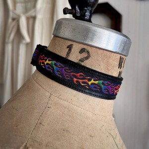 Rainbow flame print jacquard ribbon upper sewn onto a one inch wide black faux vegan leather choker collar base. Piece is adjustable and closes in back with a black metal hook. Tags lgbtq pride month queer gay goth flamer ribbon necklace punk rock