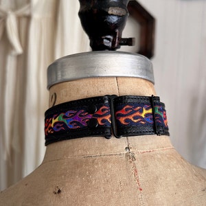 Rainbow flame print jacquard ribbon upper sewn onto a one inch wide black faux vegan leather choker collar base. Piece is adjustable and closes in back with a black metal hook. Tags lgbtq pride month queer gay goth flamer ribbon necklace punk rock