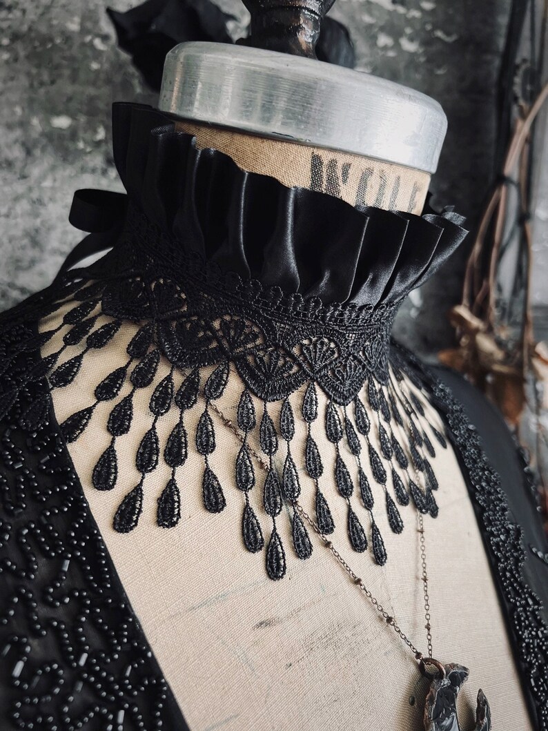 A wide black lace choker collar encircles the neck. Lace is detailed, textured and falls in dainty teardrop fringe past the collarbones. Choker is topped with wide black satin pleated trim. Tags romantic gothic dark fashion vampire wedding mourning