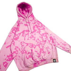 Puede incluir: Una sudadera con capucha rosa te&ntilde;ida con tinte anudado con un bolsillo de canguro y una capucha con cord&oacute;n. La sudadera presenta una base rosa claro con patrones de te&ntilde;ido anudado rosa brillante. Una peque&ntilde;a etiqueta negra con un gr&aacute;fico blanco est&aacute; en la parte inferior izquierda.