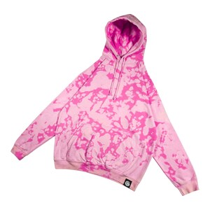Puede incluir: Una sudadera con capucha rosa con un estampado tie-dye en un tono rosa m&aacute;s oscuro. La sudadera tiene una capucha con cord&oacute;n, un bolsillo delantero y mangas largas. Un peque&ntilde;o parche con el logo es visible en la parte inferior izquierda.