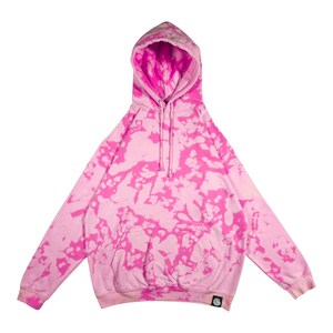 Puede incluir: Una sudadera con capucha rosa con un estampado tie-dye en rosa m&aacute;s brillante. La sudadera tiene una capucha con cord&oacute;n, un bolsillo delantero y mangas largas. Un peque&ntilde;o logotipo es visible en la parte inferior izquierda.