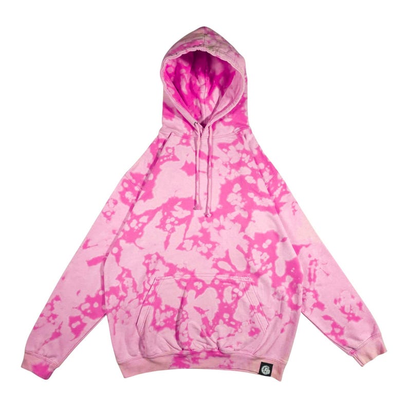 Puede incluir: Una sudadera con capucha rosa con un estampado tie-dye en rosa m&aacute;s brillante. La sudadera tiene una capucha con cord&oacute;n, un bolsillo delantero y mangas largas. Un peque&ntilde;o logotipo es visible en la parte inferior izquierda.
