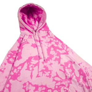 Puede incluir: Una sudadera con capucha rosa te&ntilde;ida con un cord&oacute;n y un bolsillo de canguro. La sudadera tiene una base rosa claro con vibrantes patrones de te&ntilde;ido anudado rosa. El interior de la capucha es de color rosa brillante.