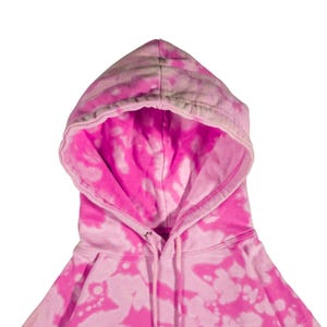 Puede incluir: Primer plano de una sudadera con capucha rosa te&ntilde;ida con un cord&oacute;n. El interior de la capucha es de un rosa vibrante, que contrasta con el patr&oacute;n de te&ntilde;ido anudado rosa claro y blanco del exterior. La sudadera tiene un dise&ntilde;o cl&aacute;sico.