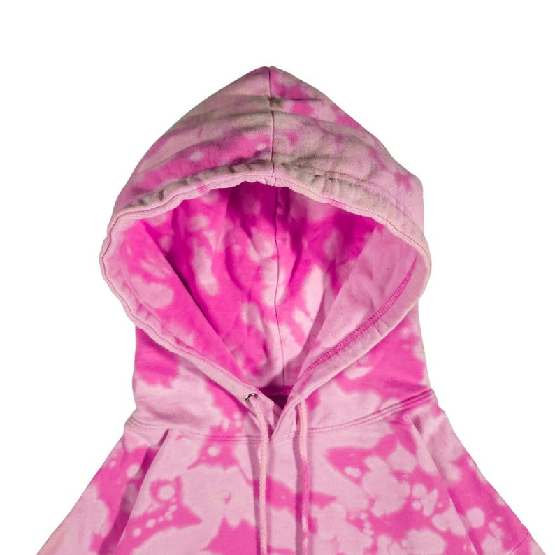 Puede incluir: Primer plano de una sudadera con capucha rosa te&ntilde;ida con un cord&oacute;n. El interior de la capucha es de un rosa vibrante, que contrasta con el patr&oacute;n de te&ntilde;ido anudado rosa claro y blanco del exterior. La sudadera tiene un dise&ntilde;o cl&aacute;sico.