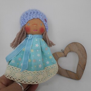 Peut inclure: Une poupée faite à la main portant une robe à motifs d'étoiles bleues et blanches, une ceinture en velours bleu et un bonnet en tricot bleu clair. La poupée a des cheveux en laine brune et un visage peint avec des joues roses.