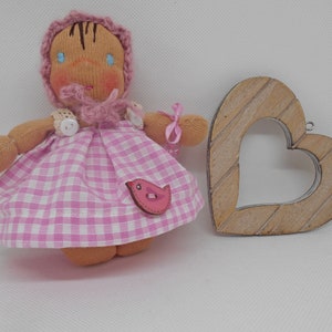 Peut inclure: Une poupée faite à la main portant une robe en vichy rose et blanche, un chapeau rose en crochet et des yeux bleus. La poupée tient un petit oiseau rose. Un cœur en bois est en arrière-plan.