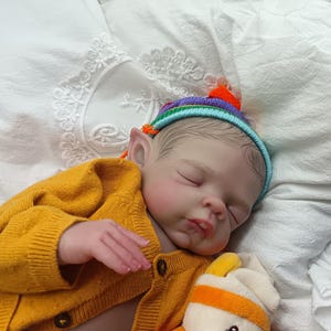 Peut inclure: Une poupée réaliste aux oreilles pointues d'elfe, vêtue d'un gilet en tricot jaune moutarde et d'un petit bonnet rayé coloré avec un pompon orange vif. La poupée a les yeux fermés, des cheveux peints et repose sur un tissu blanc.