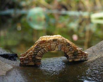 Può includere: Un piccolo ponte in miniatura di pietra, perfetto per un giardino fatato o una scena in miniatura. Il ponte è fatto di pietre marroni e beige e ha un arco arrotondato.