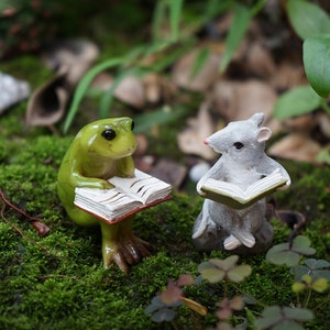 May include: A green frog and a gray mouse figurine are sitting on a bed of moss, each holding an open book. The frog is sitting on its hind legs, while the mouse is sitting on its haunches. The figurines are both looking at their books.