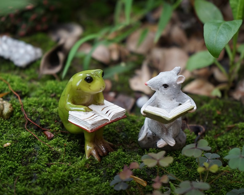 May include: A green frog and a gray mouse figurine are sitting on a bed of moss, each holding an open book. The frog is sitting on its hind legs, while the mouse is sitting on its haunches. The figurines are both looking at their books.