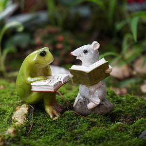 May include: A green frog and a gray mouse figurine sitting on a bed of green moss, each holding an open book. The frog is on the left and the mouse is on the right.