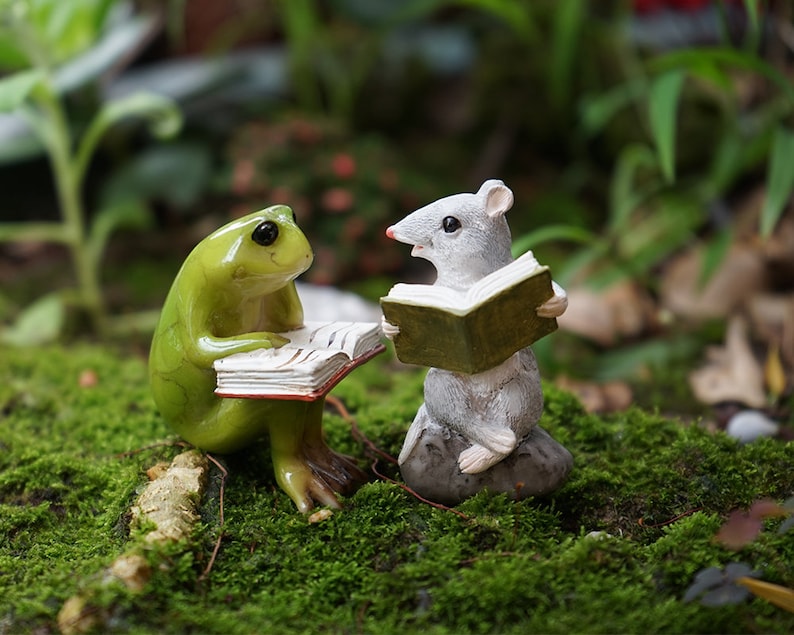May include: A green frog and a gray mouse figurine sitting on a bed of green moss, each holding an open book. The frog is on the left and the mouse is on the right.