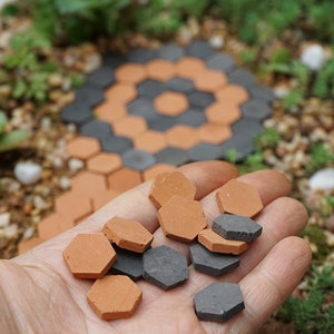 May include: A hand holding a variety of small hexagonal tiles. The tiles are made of terracotta and a dark gray material. The tiles are arranged in a pattern, with the terracotta tiles on top of the gray tiles.