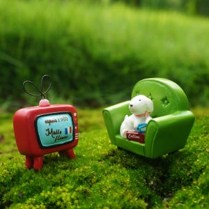 May include: A miniature red and white television with the text "expo 1986" and "Malle Fleurs" on the screen. A miniature green armchair with a white dog figurine sitting on it. The dog is holding a book with the text "Collins" on the cover.
